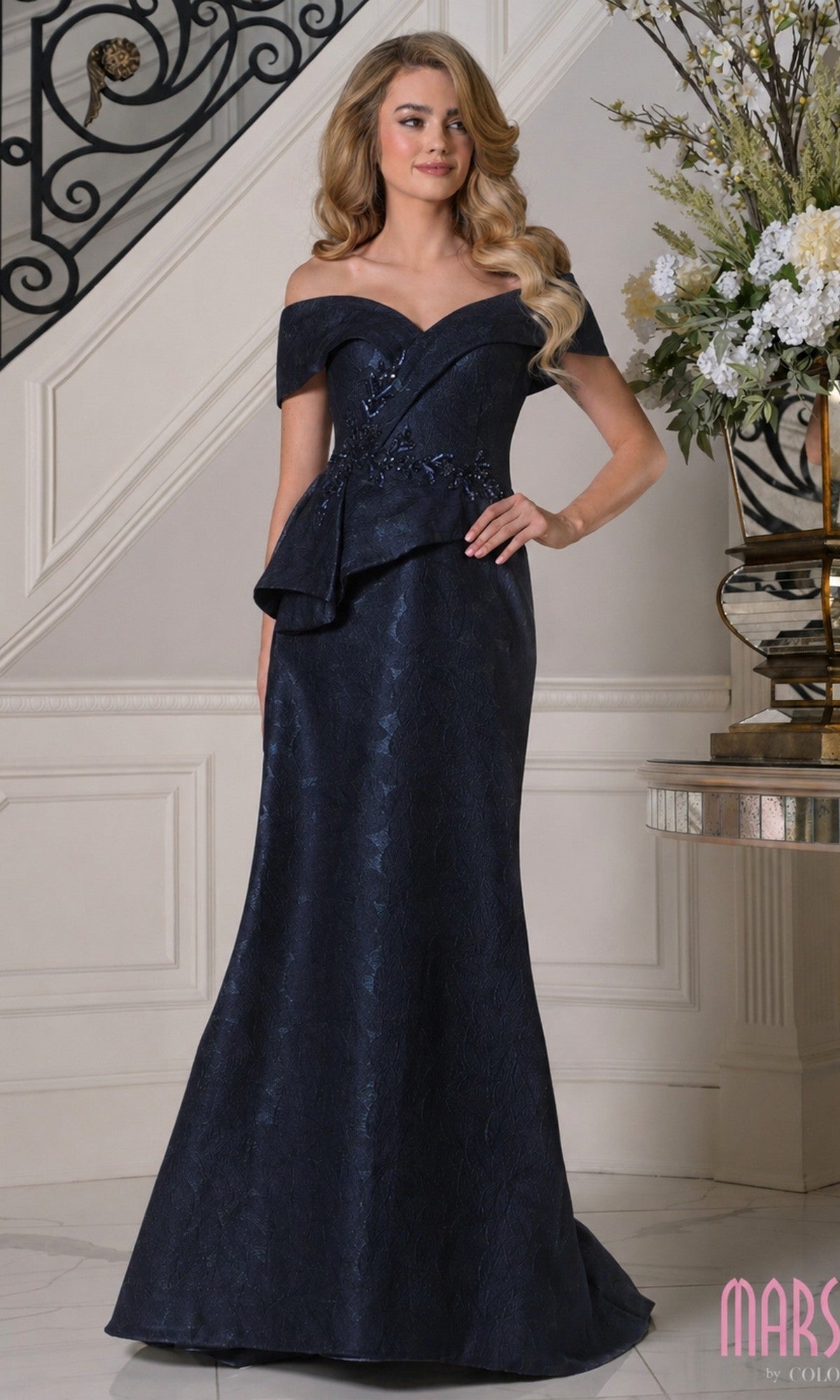 A woman stands indoors wearing the Marsoni MV1397 Long Formal Dress in navy, featuring an off-the-shoulder design, floral jacquard details, and an asymmetrical peplum waist. She poses by a staircase and elegant floral arrangement.